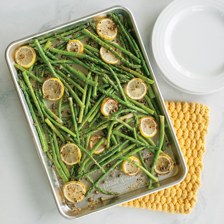 Roasted green beans with lemon slices on a baking tray, placed on a textured yellow mat.