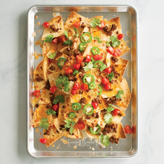 Metal tray with loaded nachos garnished with tomatoes, jalapenos, and cilantro on a light gray background.