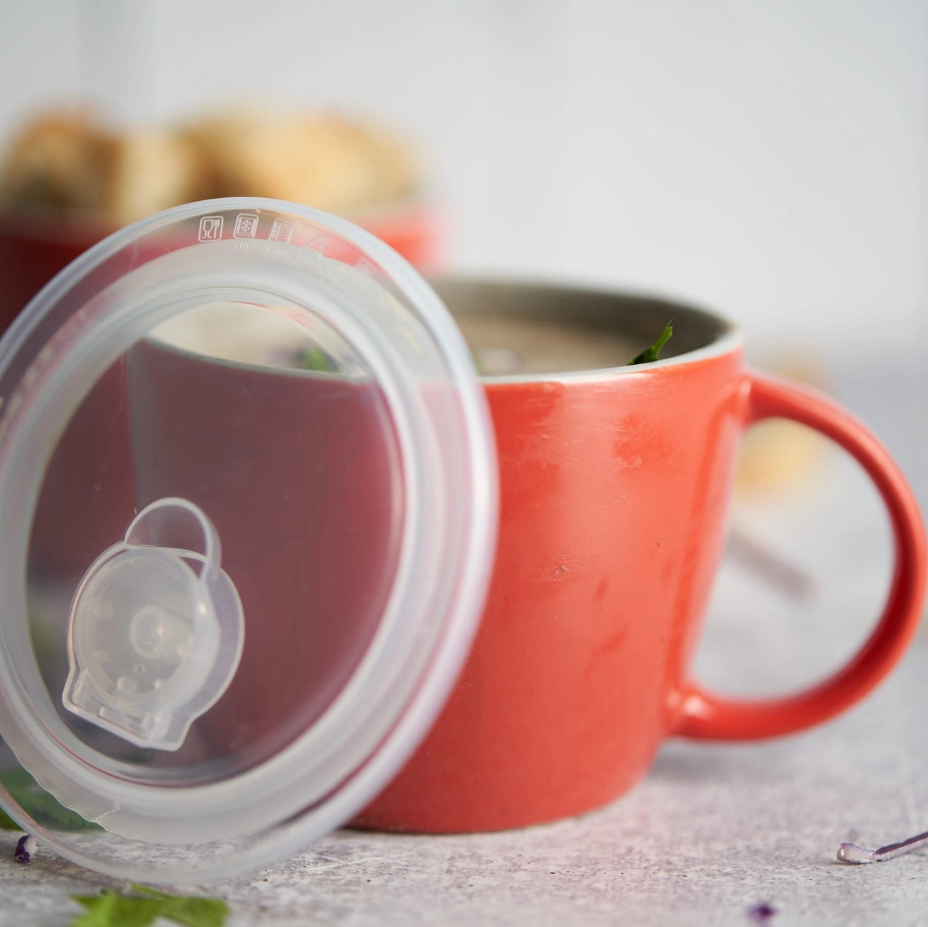 A clear silicone lid with a small clear plastic strainer or filter piece, shown leaning against an orange-red ceramic mug on a light tabletop.