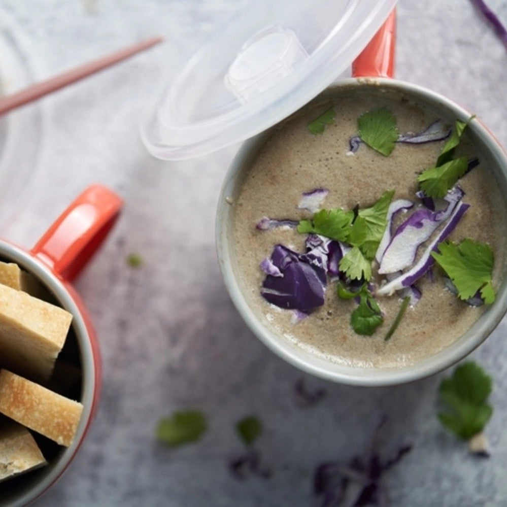 A styled overhead shot of a creamy mushroom soup in a gray mug with a red handle, garnished with cilantro and purple cabbage, served alongside cubed bread in another mug and a slice of bread on a plate.