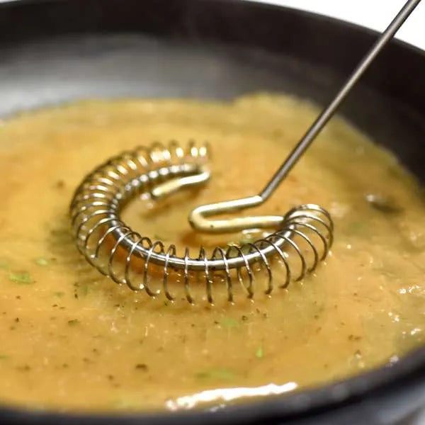 A close-up of a metal whisk with a coiled  body and a straight handle, resting in a bowl of yellow-orange liquid .