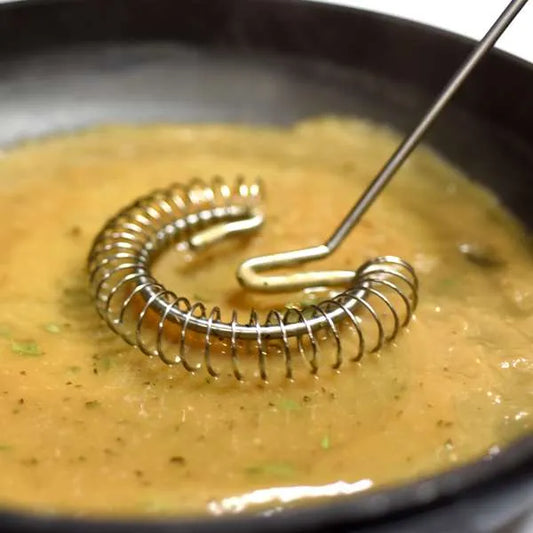 A close-up of a metal whisk with a coiled  body and a straight handle, resting in a bowl of yellow-orange liquid .