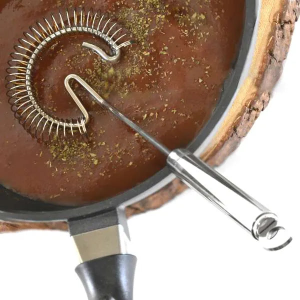 A stainless steel spiral whisk with a long handle, shown resting inside a black frying pan with a brown sauce, on a white background.