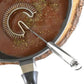 A stainless steel spiral whisk with a long handle, shown resting inside a black frying pan with a brown sauce, on a white background.