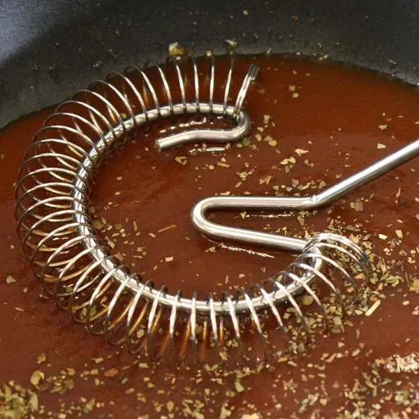 A stainless steel spiral whisk or emulsifier resting in a pan with reddish-brown liquid and scattered herbs.