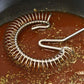 A stainless steel spiral whisk or emulsifier resting in a pan with reddish-brown liquid and scattered herbs.