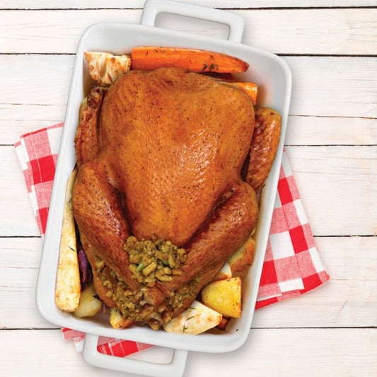 A roasted whole turkey in a white rectangular baking dish with sides, surrounded by carrots, potatoes, and stuffing, placed on a red-and-white checkered cloth over a white wood plank surface.