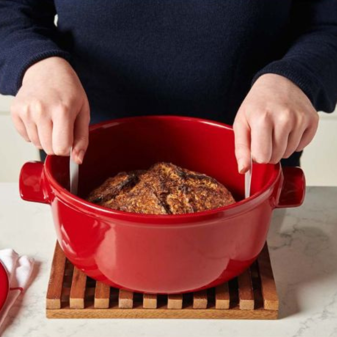 Hands using a white loaf lifter to begin to pull a loaf of bread out of a red dutch oven