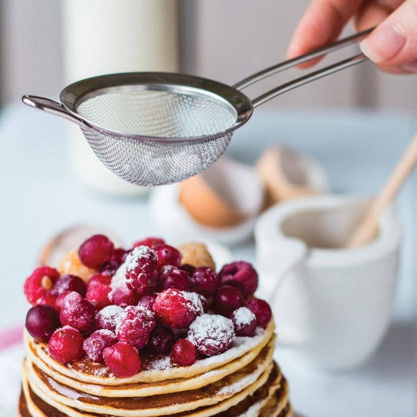 Stack of pancakes with berries being dusted with powdered sugar using a small sieve.