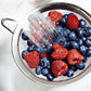 Berries being rinsed under running water in a metal strainer.