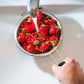Strawberries being washed under running water in a colander over a sink.