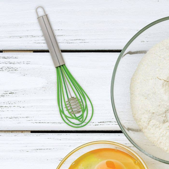 Green whisk on a white wooden surface with a glass bowl of dough and a small bowl of liquid.