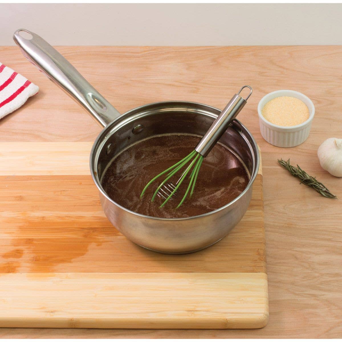 Stainless steel saucepan with whisk on a wooden cutting board, ingredients in the background