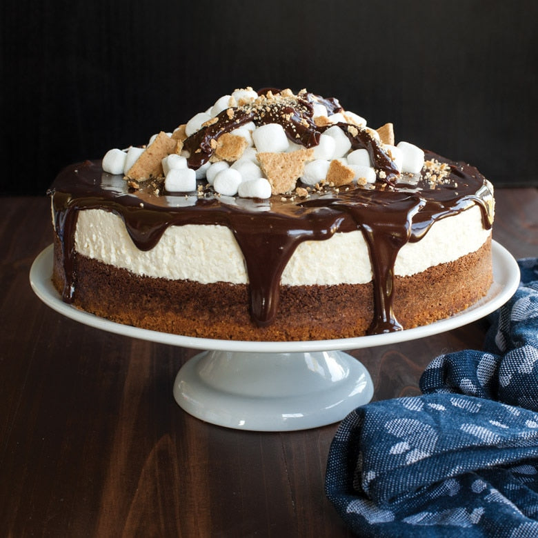 S'mores cheesecake with chocolate drizzle on a white cake stand against a dark background