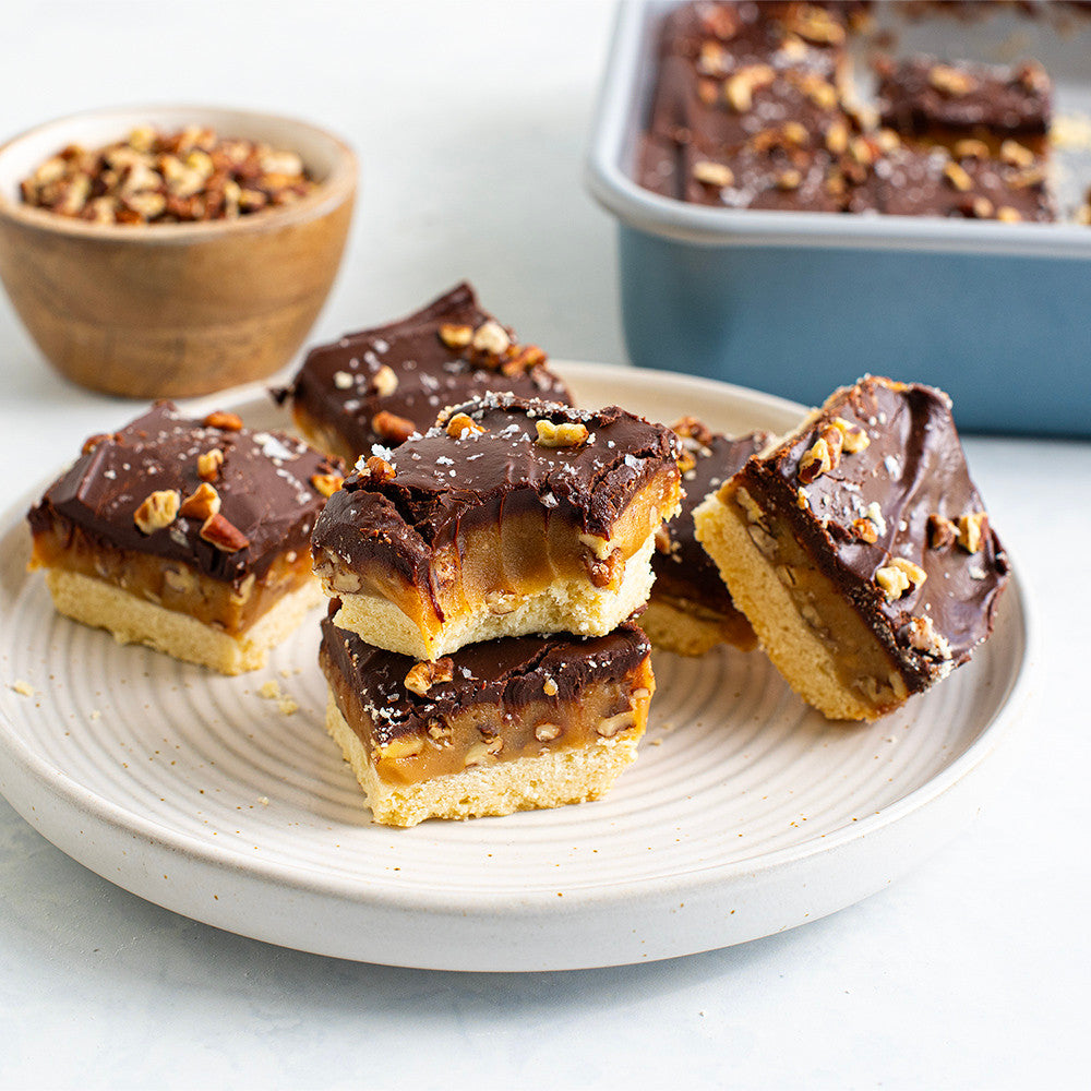 Chocolate-covered caramel bars on a plate with a bowl of nuts in the background