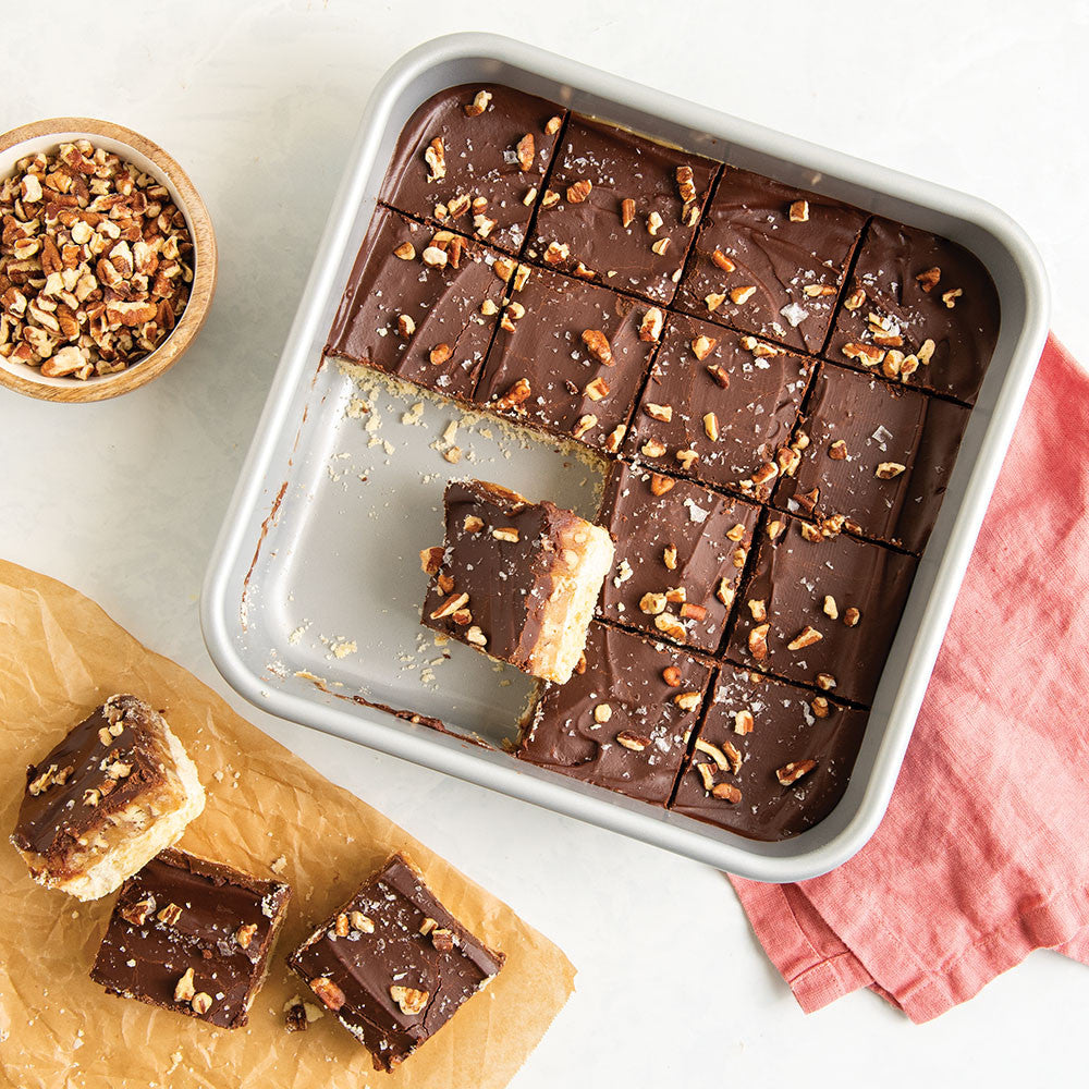 Chocolate dessert bars in a pan with a bowl of nuts on a light surface.