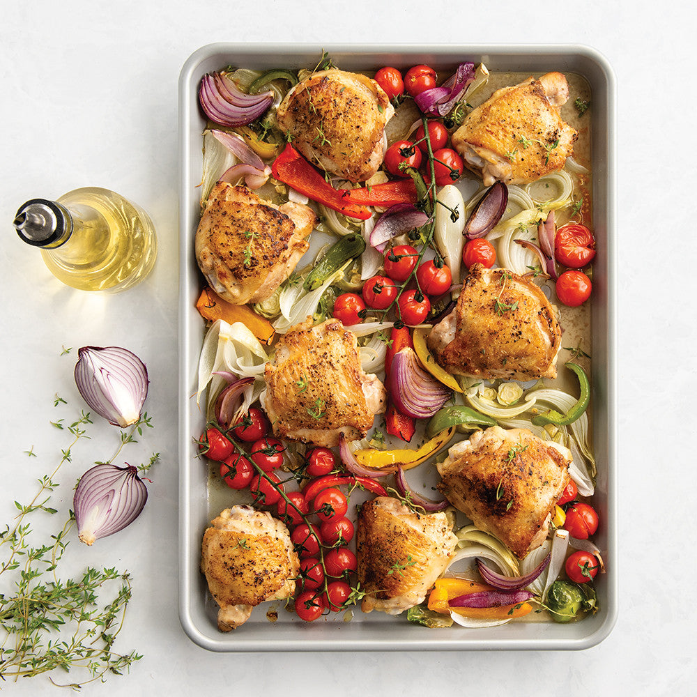 Sheet pan with roasted chicken and vegetables on a white surface with herbs and oil.