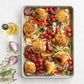 Sheet pan with roasted chicken and vegetables on a white surface with herbs and oil.