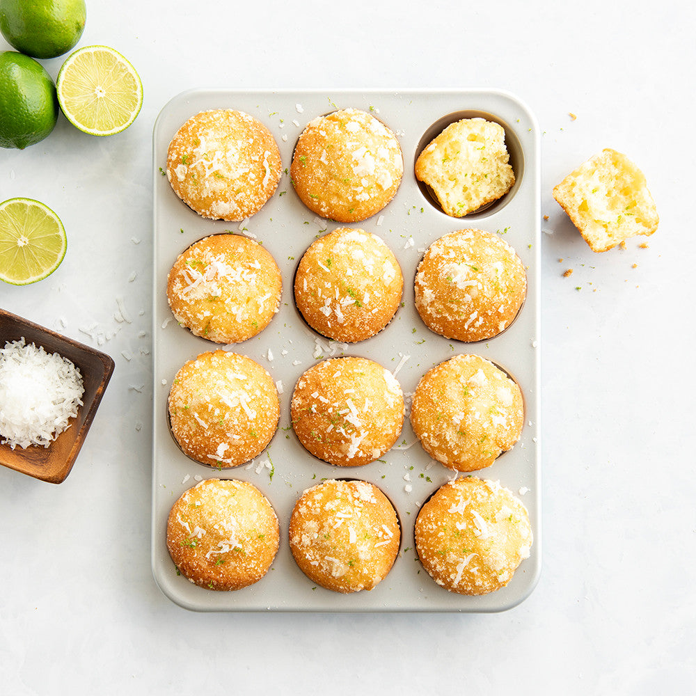 Muffin tin with coconut muffins on a white surface with limes and coconut flakes.