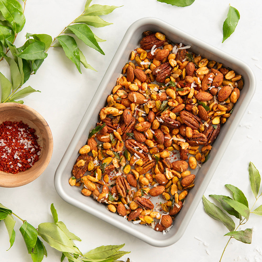 Tray of roasted mixed nuts with a small bowl of seasoning on a white background