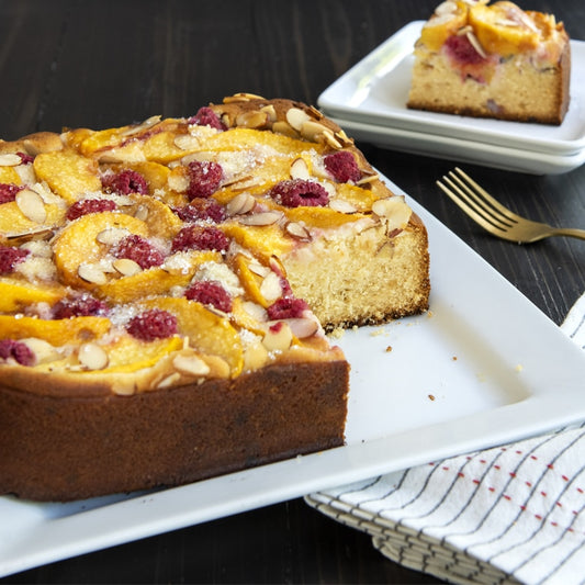 Peach and raspberry cake on a white plate with a slice removed, on a dark surface.