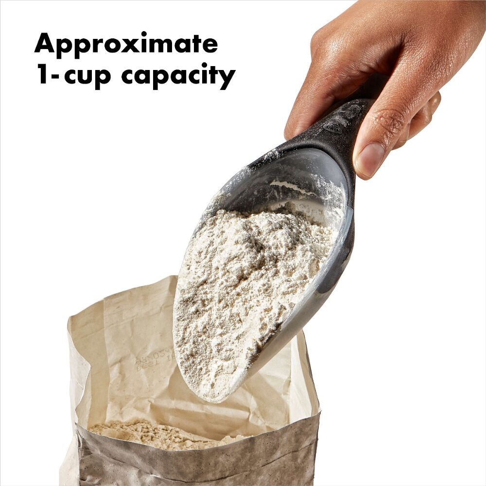 Hand using a black measuring scoop to pour flour into a paper bag with text indicating approximate 1-cup capacity.