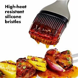 Barbecue brush with high-heat resistant silicone bristles applying sauce to grilled vegetables on a white background.
