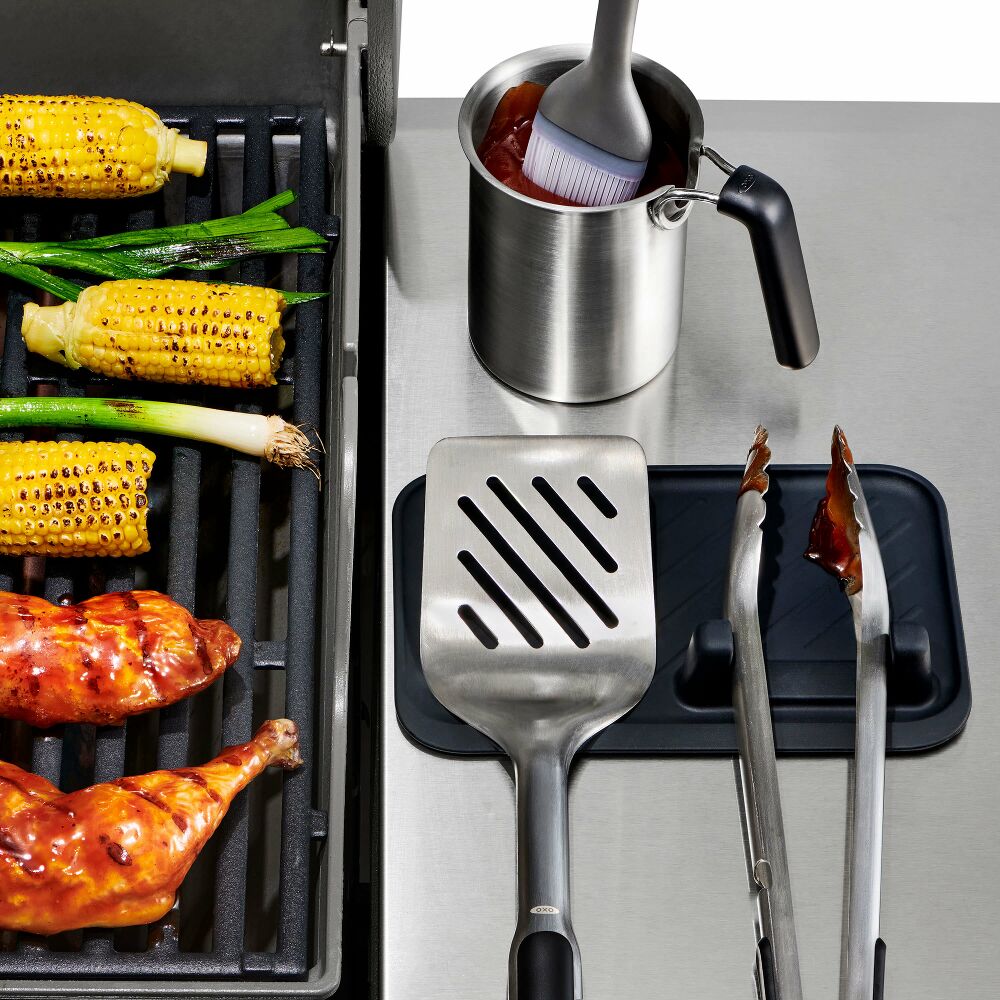 Grilling scene with corn, chicken, and cooking utensils on a metal surface.