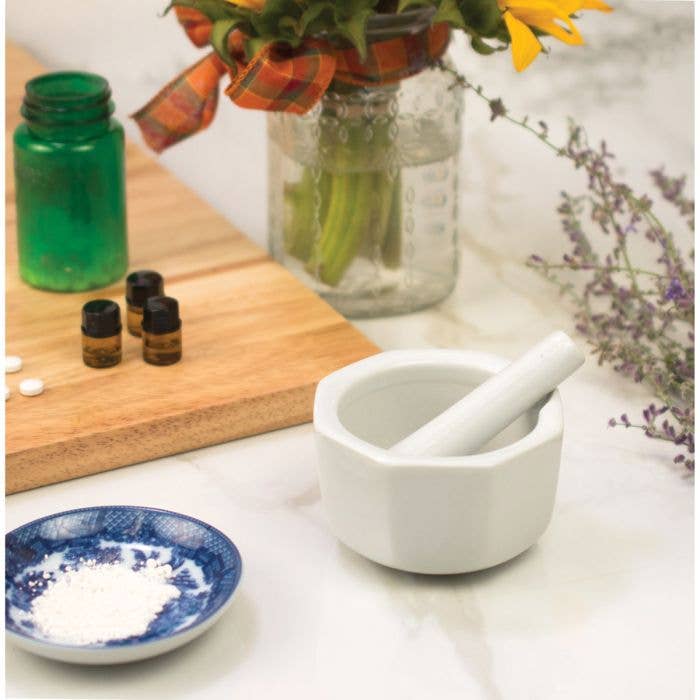 Mortar and pestle on a white surface with bottles, a cutting board, and flowers in the background.