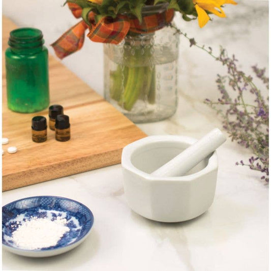 Mortar and pestle on a white surface with bottles, a cutting board, and flowers in the background.
