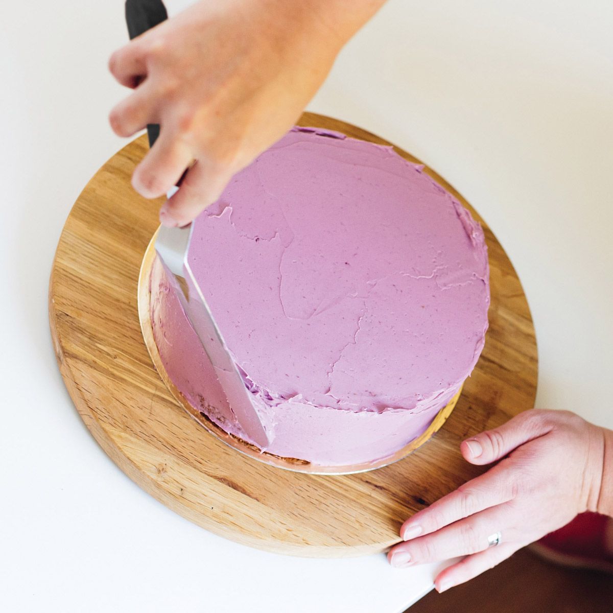Person icing a pink cake on a wooden board with a white background