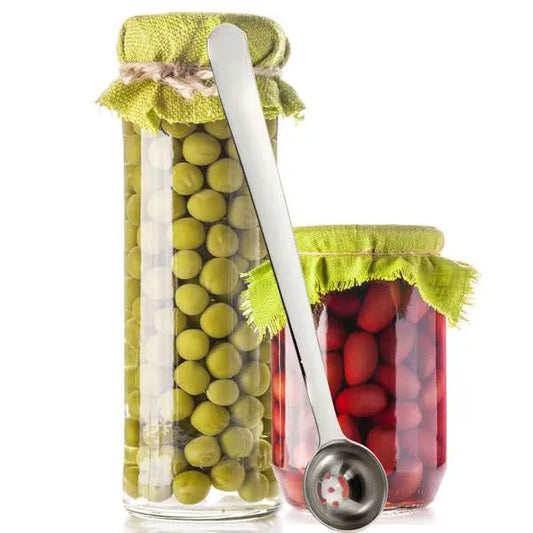 Two clear glass jars filled with green olives and red olives, topped with green cloth lids and a metal olive spoon resting across them, on a white background.