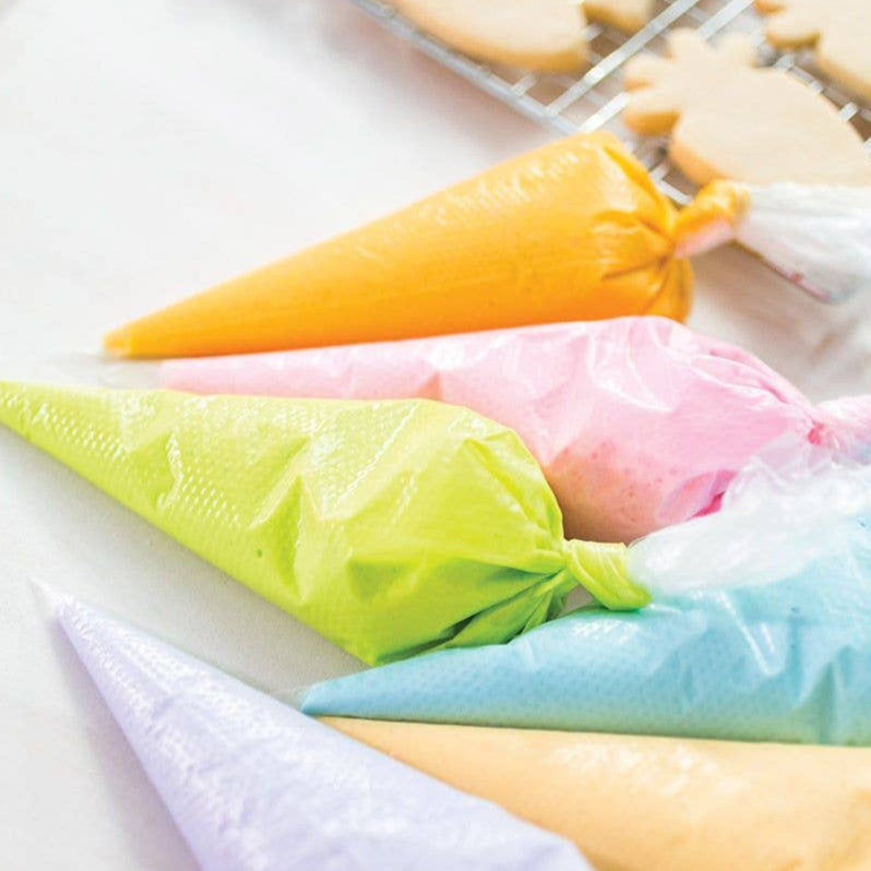 Colorful piping bags filled with icing on a white surface with cookies in the background.