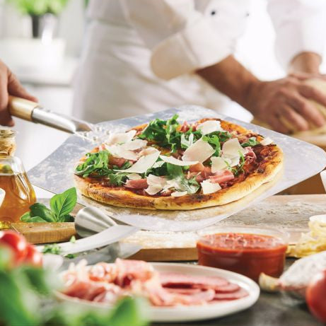 A freshly topped pizza sits on a metal pizza peel in a kitchen setting, with a blurred chef in white clothing in the background and ingredients like prosciutto, basil, and tomato visible in the foreground.