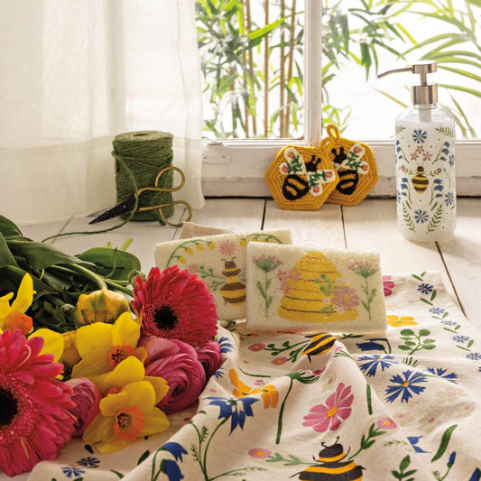 Decorative tablecloth with floral and bee patterns on a table with flowers and a window in the background.
