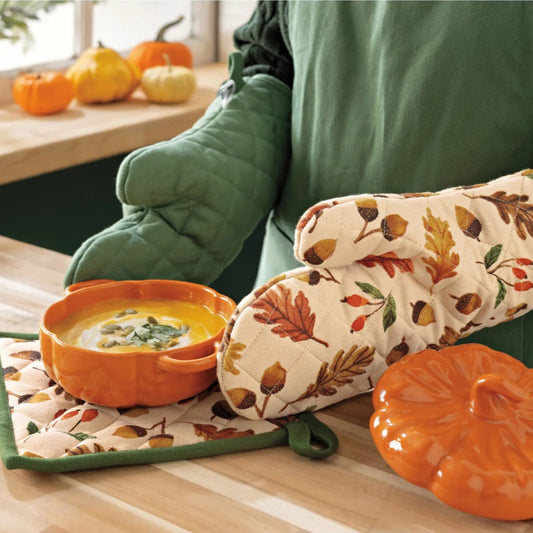 Person holding a bowl of soup with fall-themed oven mitts and pot holder on a table with pumpkins and leaves.