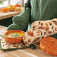 Person holding a bowl of soup with fall-themed oven mitts and pot holder on a table with pumpkins and leaves.