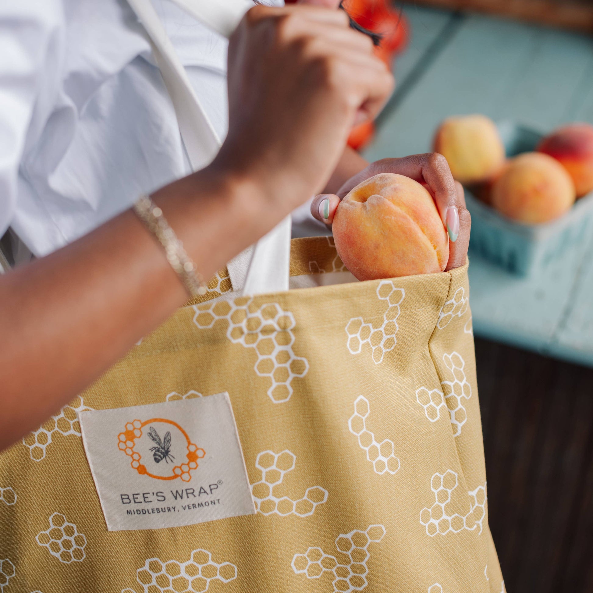 Person holding a yellow reusable bag with a brand label, filled with fruits, next to a table with more fruits.