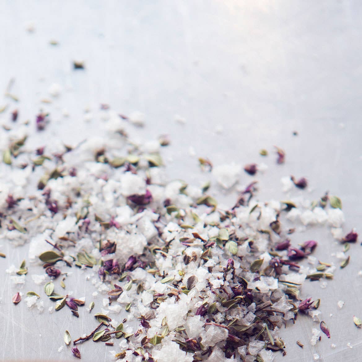 Coarse sea salt crystals mixed with dried purple flower buds and green herb pieces, scattered on a light gray surface.