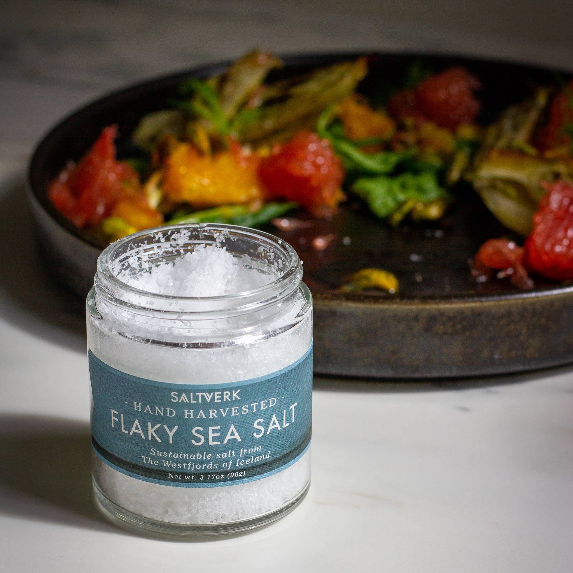 Jar of Saltwerk Flaky Sea Salt with a plate of salad in the background