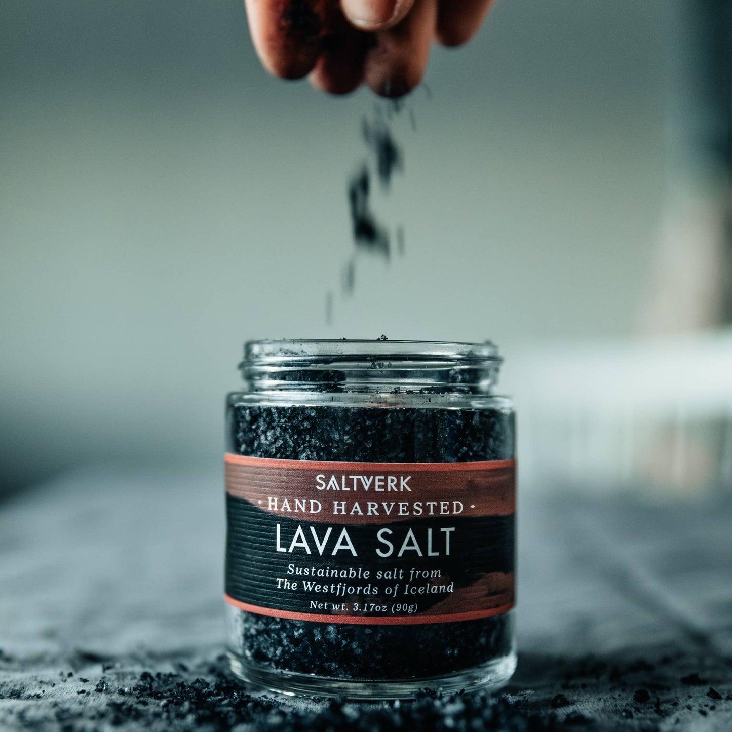 Jar of Saltwerk Lava Salt with hand sprinkling salt over it