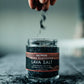 Jar of Saltwerk Lava Salt with hand sprinkling salt over it
