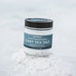 A clear glass jar of Saltyerk Flaky Sea Salt with a black lid and a dark blue label, set on white sea salt crystals against a soft, light gray background.