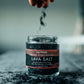 A hand sprinkles dark lava salt into a clear glass jar labeled 'SALTVERK Hand Harvested Lava Salt' on a dark textured surface.