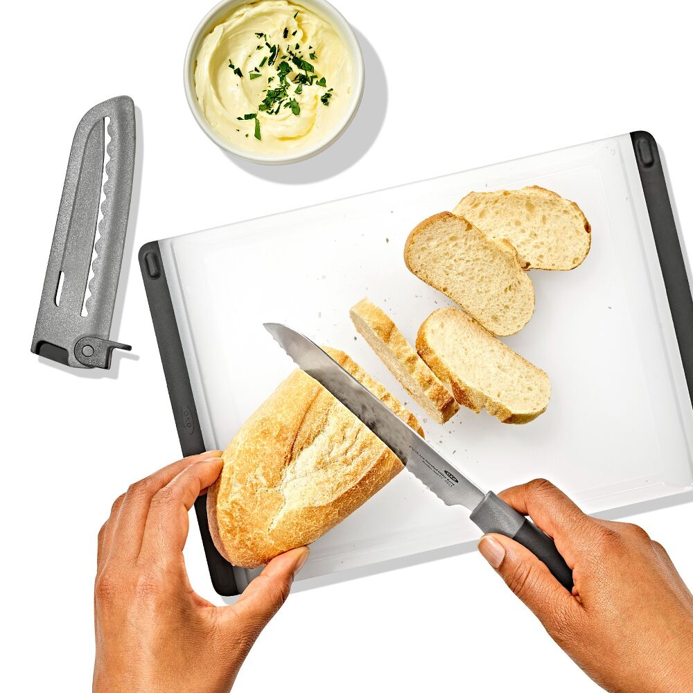 Hands using a knife to cut bread on a white cutting board. 