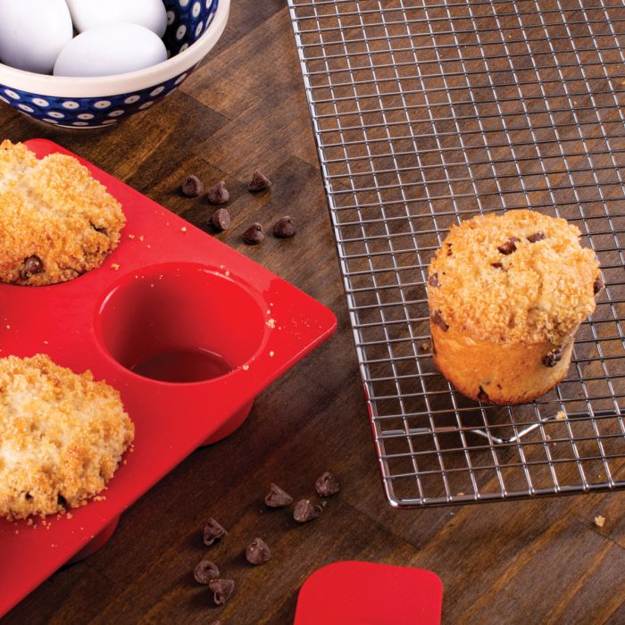 Muffins on a cooling rack with a red silicone muffin tray and bowl of eggs on a wooden surface.