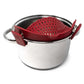 A red silicone pot strainer with side clips, shown nested inside a stainless steel pot on a white background.