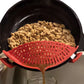 A red silicone leaf-shaped strainer held over a black pan, draining a brown liquid from cooked ground meat.