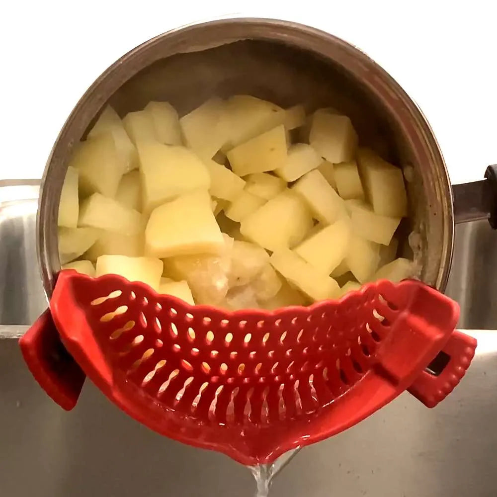 A red silicone pot strainer basket held over a metal pot filled with chopped potatoes, draining water into a sink.