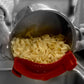 A stainless steel pot filled with uncooked egg noodles, held over a sink with a red silicone strainer basket inside the pot. The background is desaturated and blurred, with gloved hands visible on the sides.